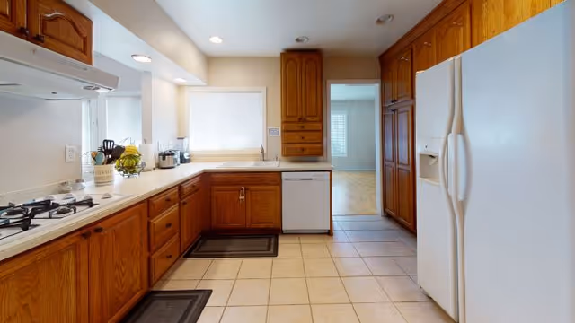 A spacious kitchen with wooden cabinets, white countertops, a white refrigerator, a dishwasher, a gas stove, and tiled flooring. There are two black floor mats in front of the sink and stove. A window with a white blind is above the sink, and the kitchen opens into another room with wooden flooring.