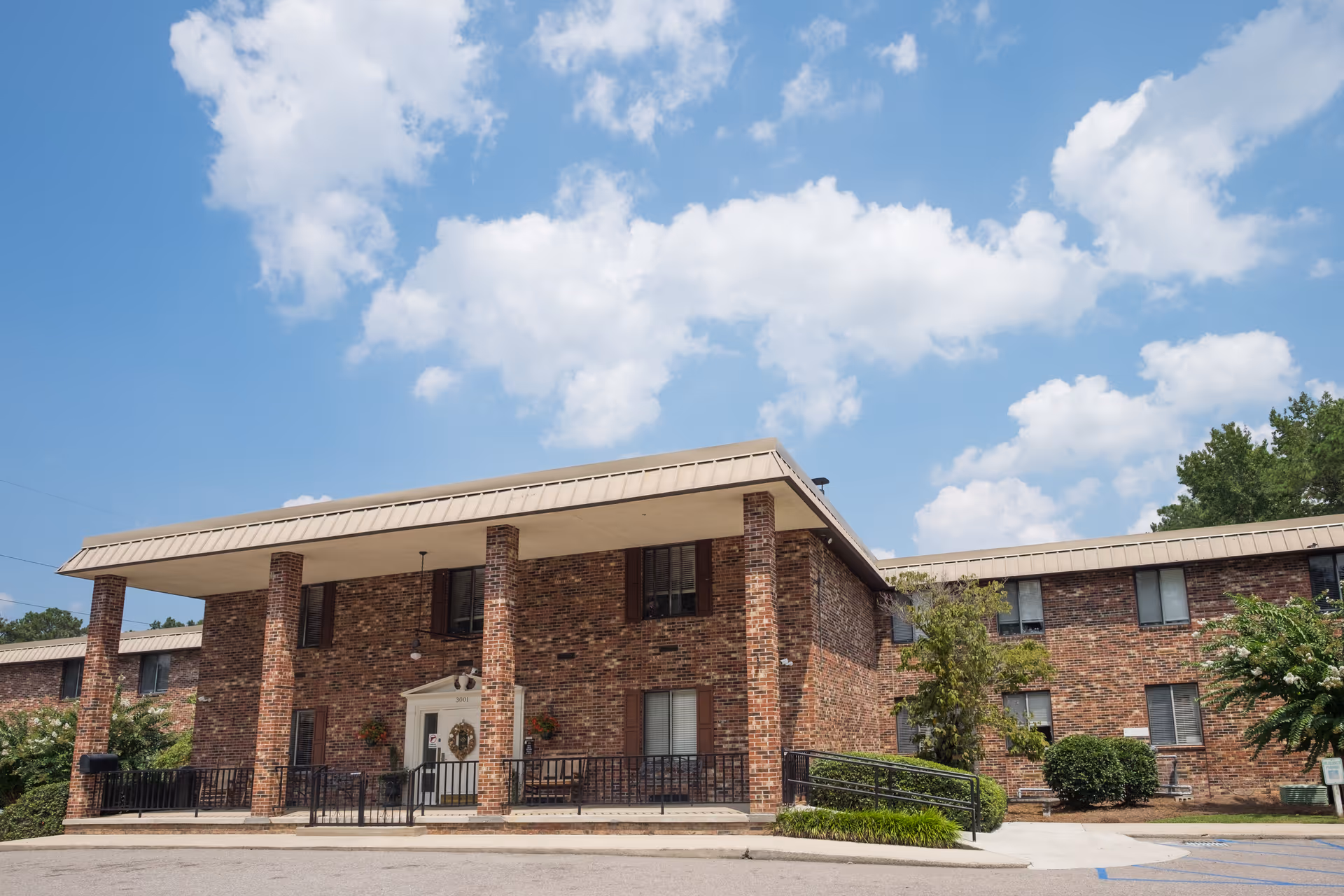 Front exterior of a two-story brick senior living building with a covered entrance, columns and a wheelchair ramp under a blue sky.