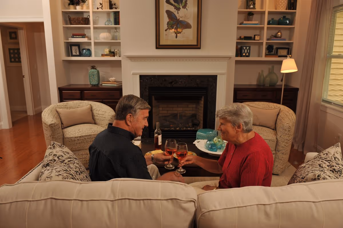 An elderly man and woman sitting on a beige couch in a cozy living room, clinking glasses of red wine in front of a fireplace. The room features two patterned armchairs, built-in shelves with decorative items, and a window with curtains.