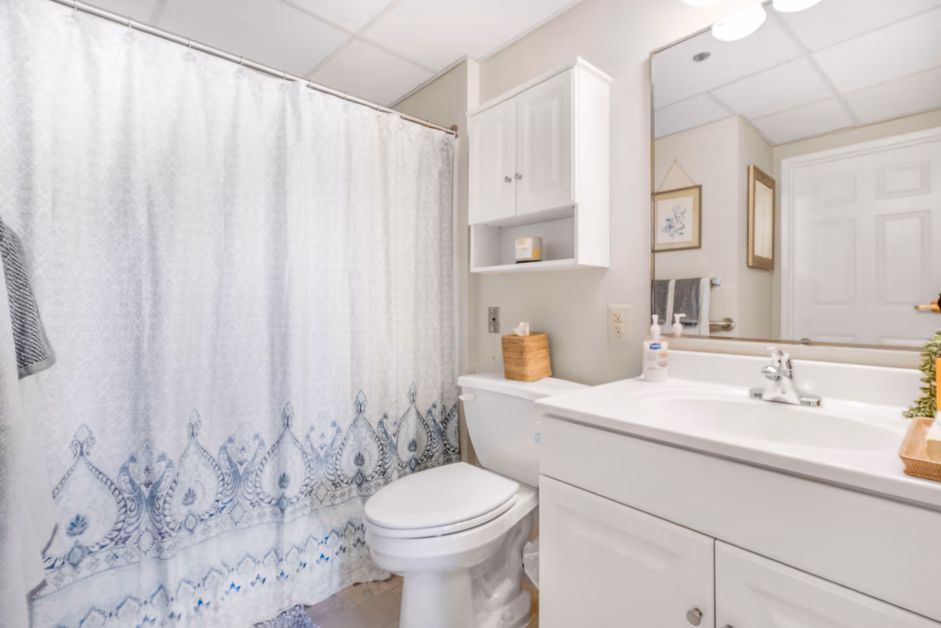 A clean and bright bathroom featuring a white toilet, a white vanity with a sink, a large mirror above the sink, a white cabinet mounted on the wall, and a shower with a white curtain decorated with a blue pattern along the bottom edge.