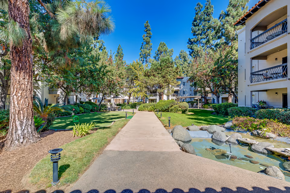 A sunny outdoor courtyard area at Solstice Senior Living at El Cajon featuring a paved walkway flanked by green grass, trees, bushes, a small pond with rocks and a water feature on the right, and apartment buildings with balconies on both sides.