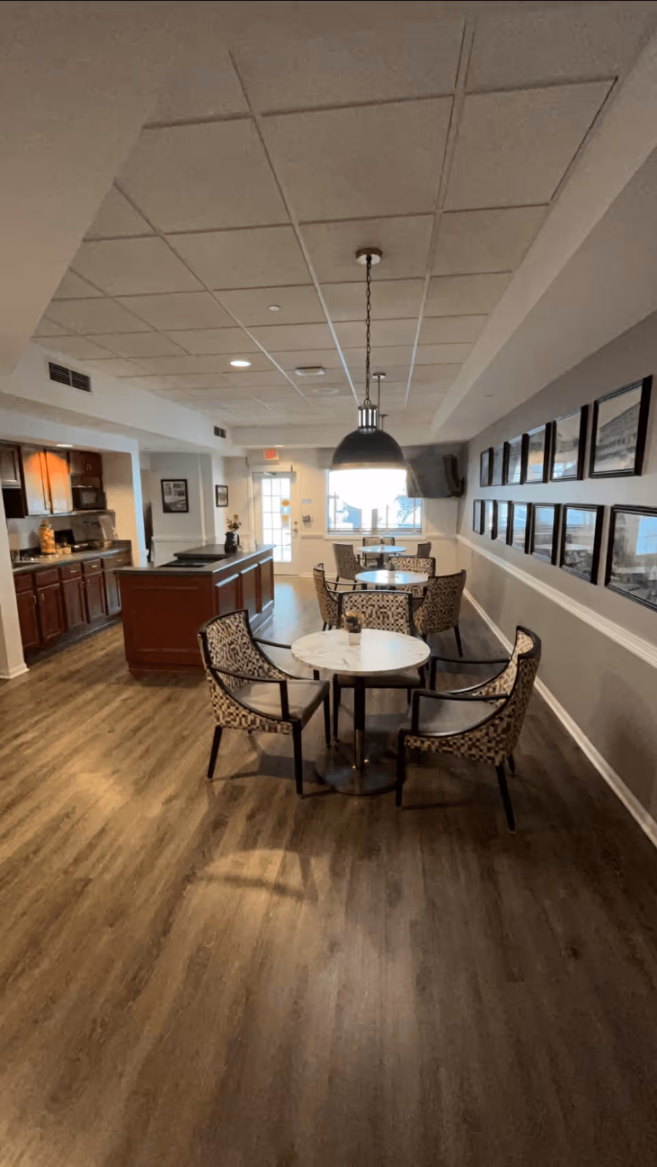Interior view of a senior living facility dining area with round tables and patterned chairs. The room features wood flooring, a kitchen area with dark wood cabinets on the left, and framed pictures on the right wall. A large pendant light hangs over one of the tables, and a door with windows is visible at the far end of the room.