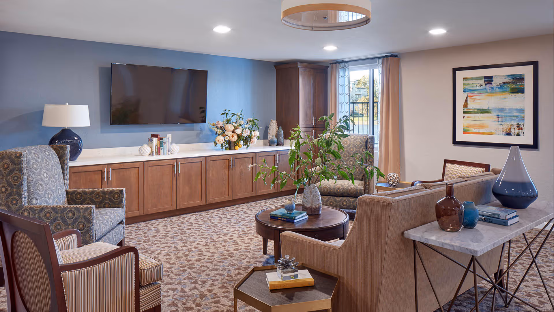 A cozy living room area with patterned armchairs, a beige sofa, a round wooden coffee table with decorative items and plants, a long wooden cabinet with books and flowers underneath a wall-mounted flat screen TV, a large window with curtains, and a framed abstract painting on the wall.