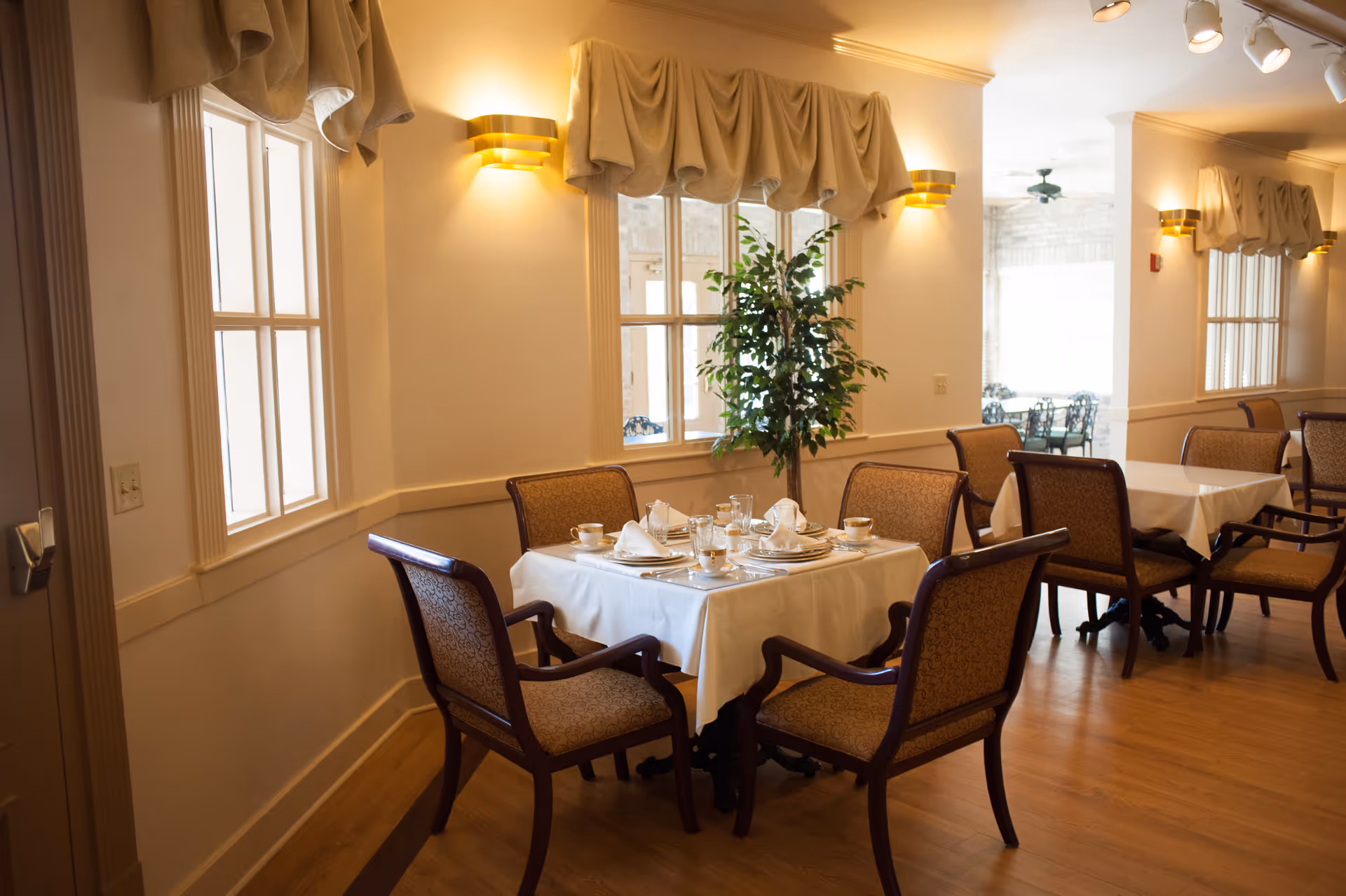 A dining room with tables covered in white tablecloths, set with cups, saucers, and napkins. The room has wooden chairs with cushioned seats and backs, windows with beige valances, wall-mounted lights, and a potted plant near the windows.
