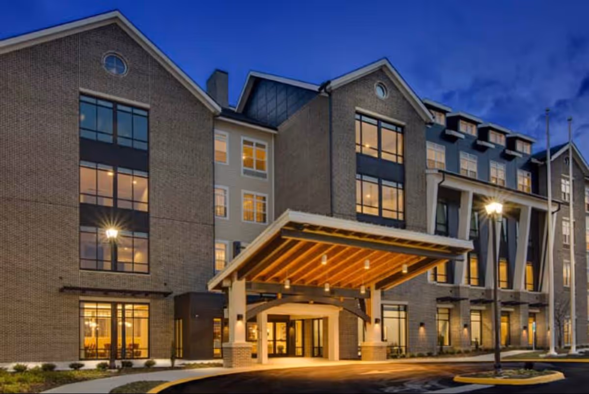 Exterior view of a multi-story senior living facility building at dusk with large windows illuminated from inside, a covered entrance with wooden beams, and a street lamp in front.
