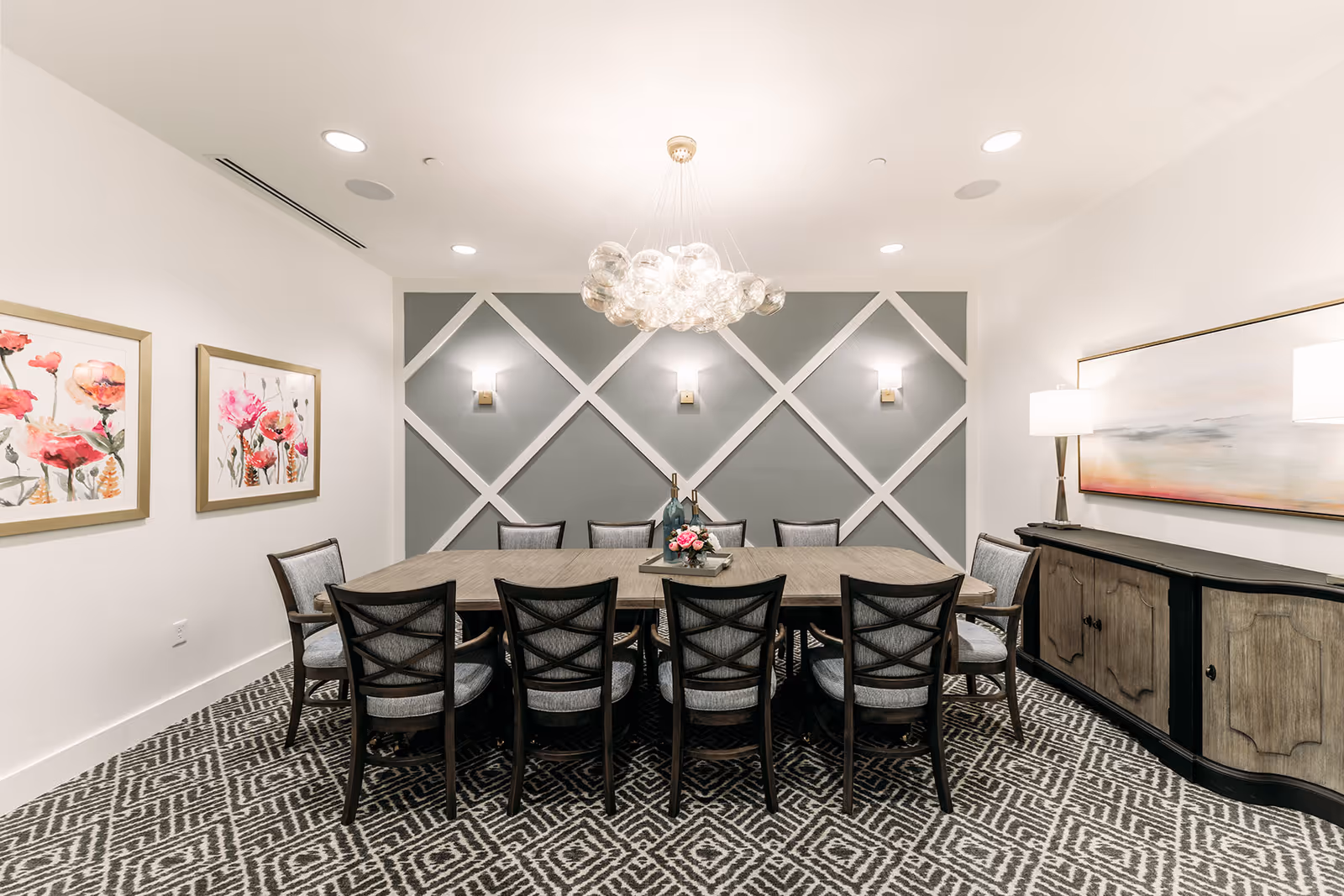 A modern dining room with a long rectangular wooden table surrounded by ten cushioned chairs. The back wall features a geometric design with white trim on a gray background and three wall sconces. A contemporary chandelier with multiple glass globes hangs above the table. The room has patterned carpet flooring, two framed floral paintings on the left wall, and a sideboard with a large abstract painting and two lamps on the right.