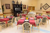 Communal dining room with round tables covered in red tablecloths and set with plates, surrounded by floral-upholstered chairs, with a piano and cabinet against the wall.