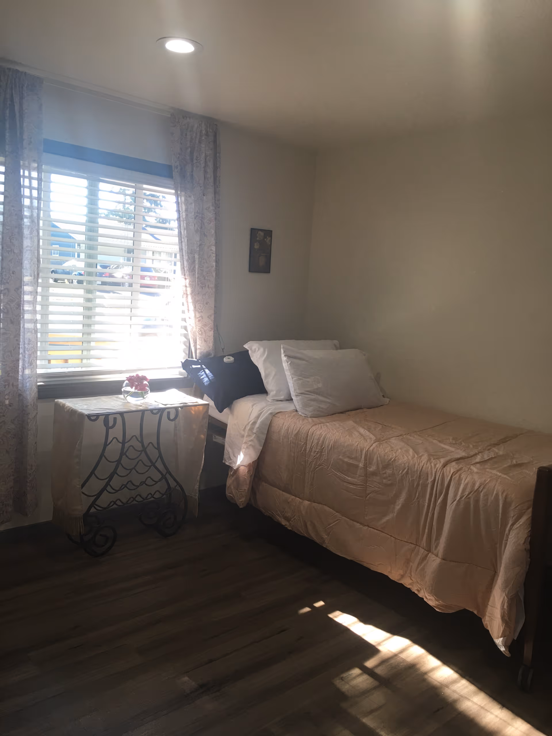 A small bedroom with a single bed covered in a beige comforter and white pillows. Next to the bed is a decorative metal table with a light-colored cloth and a small vase with flowers. A window with white blinds and patterned curtains lets in natural light, illuminating the wooden floor.