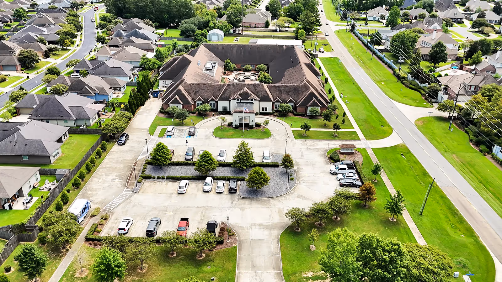 Aerial view of The Pearl at Jamestown - Memory Care Community, showing a large U-shaped building with a central courtyard, surrounded by parking lots and green lawns. The facility is located in a suburban neighborhood with houses and streets visible around it.
