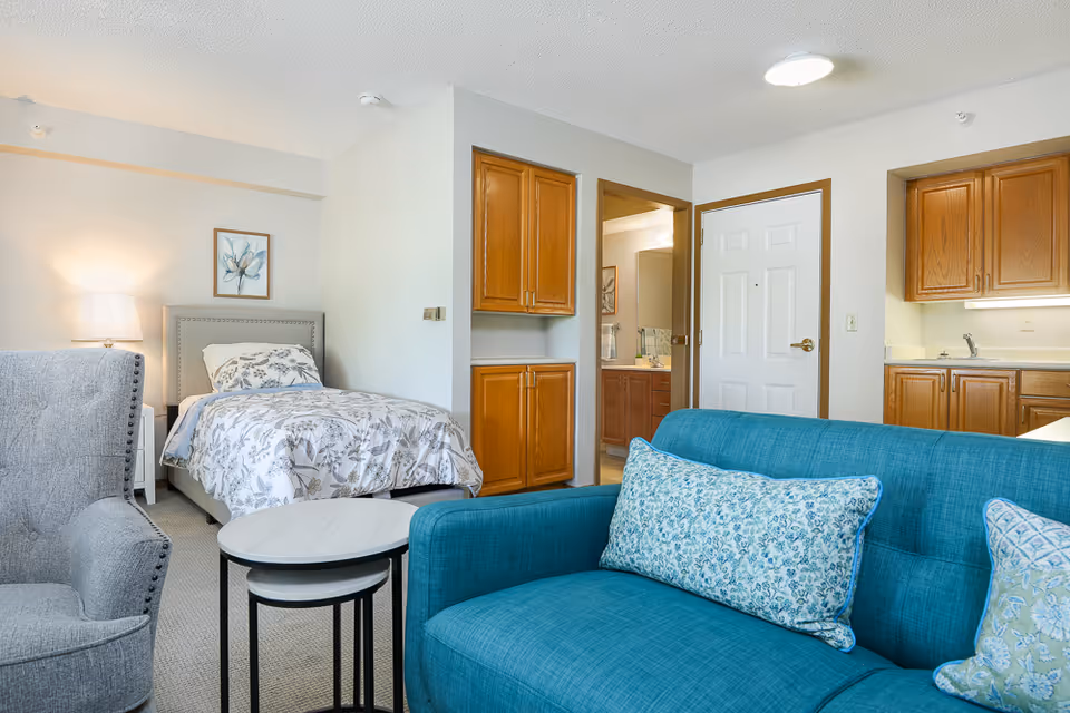 Bright studio-style assisted living apartment with a bed, teal sofa, armchair, kitchenette, and bathroom door.