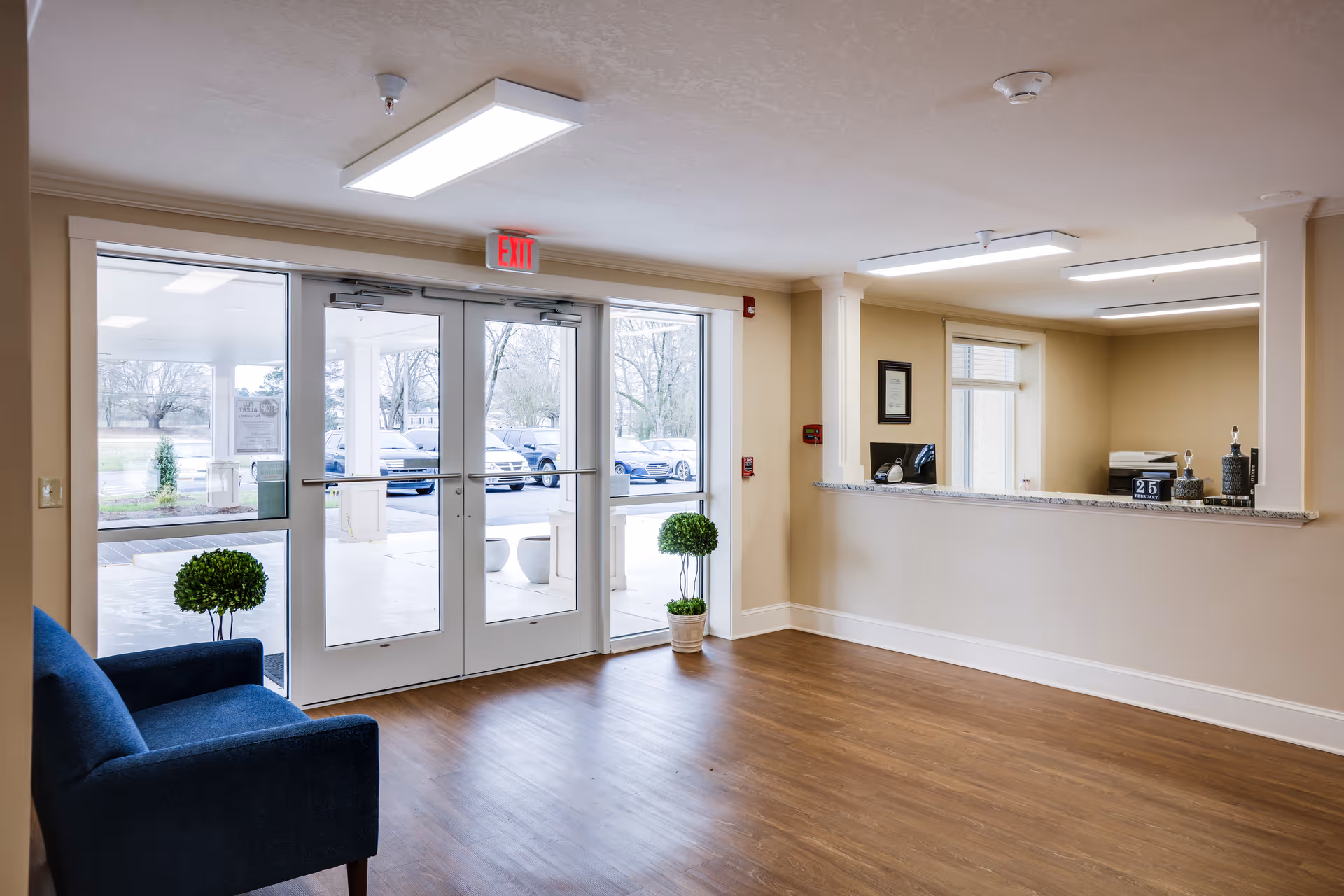 Interior view of a senior living facility entrance area with double glass doors leading outside. The room has wood flooring, beige walls, a blue armchair on the left, two small potted plants near the doors, and a reception desk with a granite countertop on the right. Fluorescent ceiling lights illuminate the space.