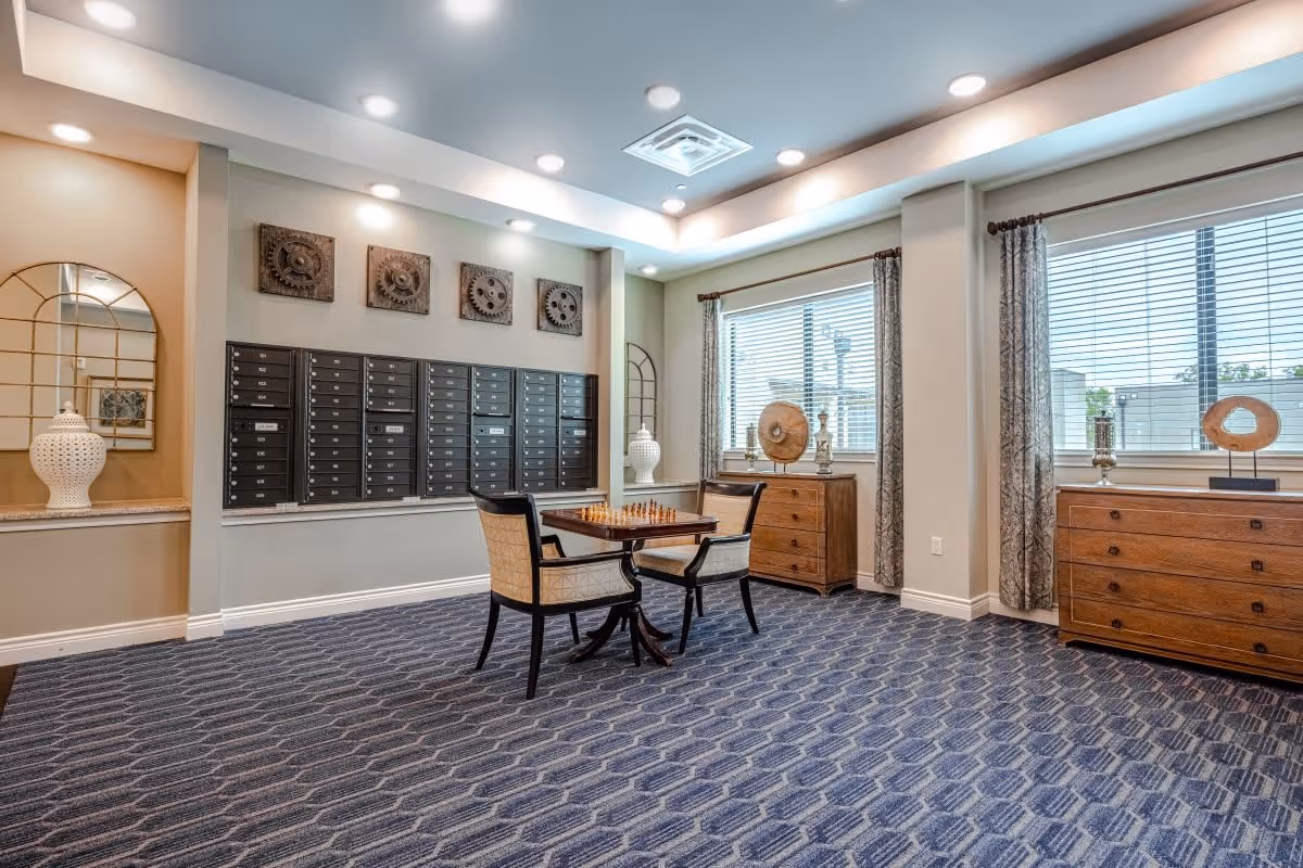A well-lit interior room with a patterned blue carpet, two wooden dressers under large windows with blinds and curtains, a small table with a chessboard and two chairs, and a wall of black mailboxes. Decorative items and wall art are also visible.