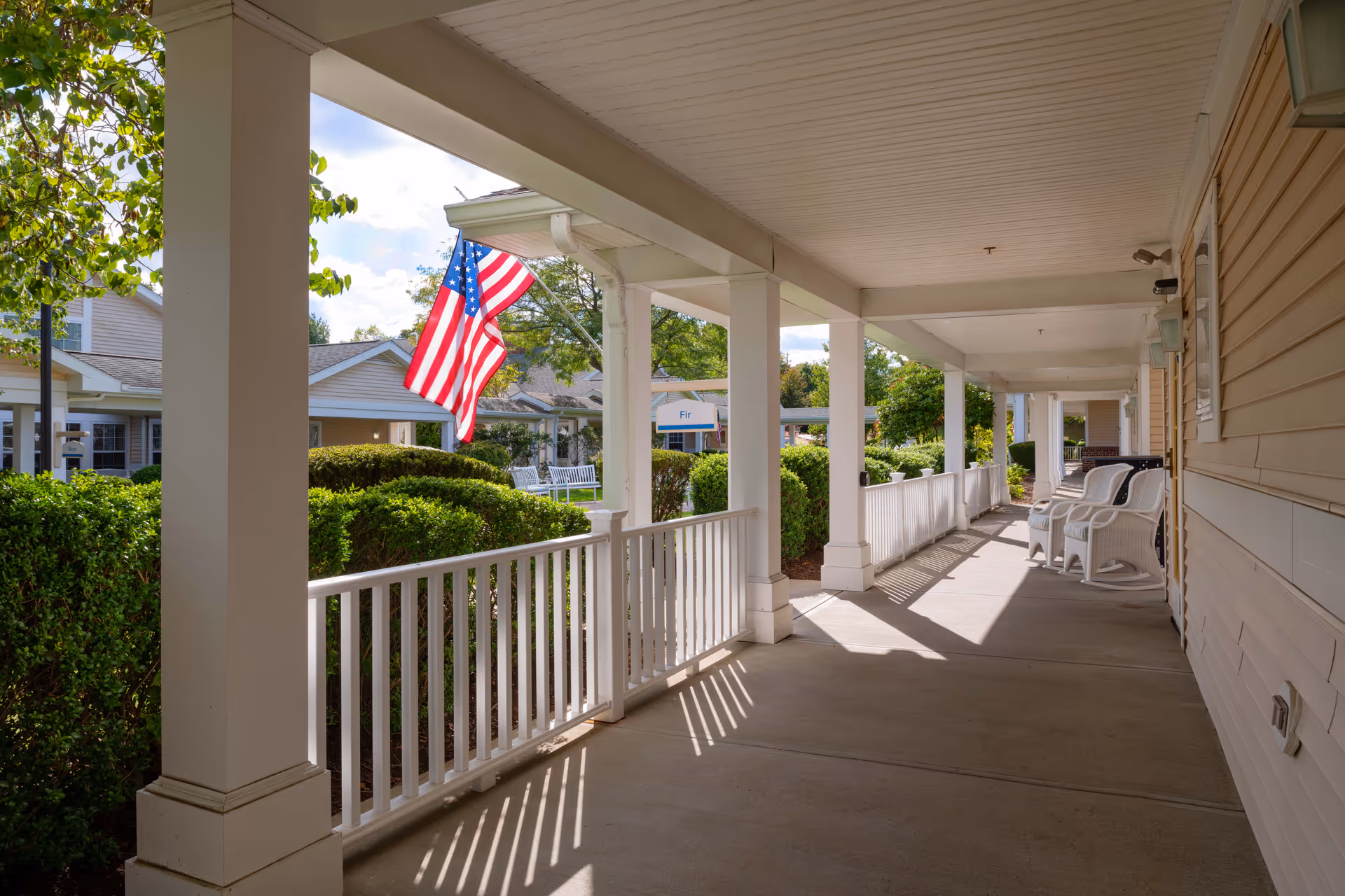 Covered porch walkway with white columns and railings, plastic chairs, an American flag, and manicured shrubs.