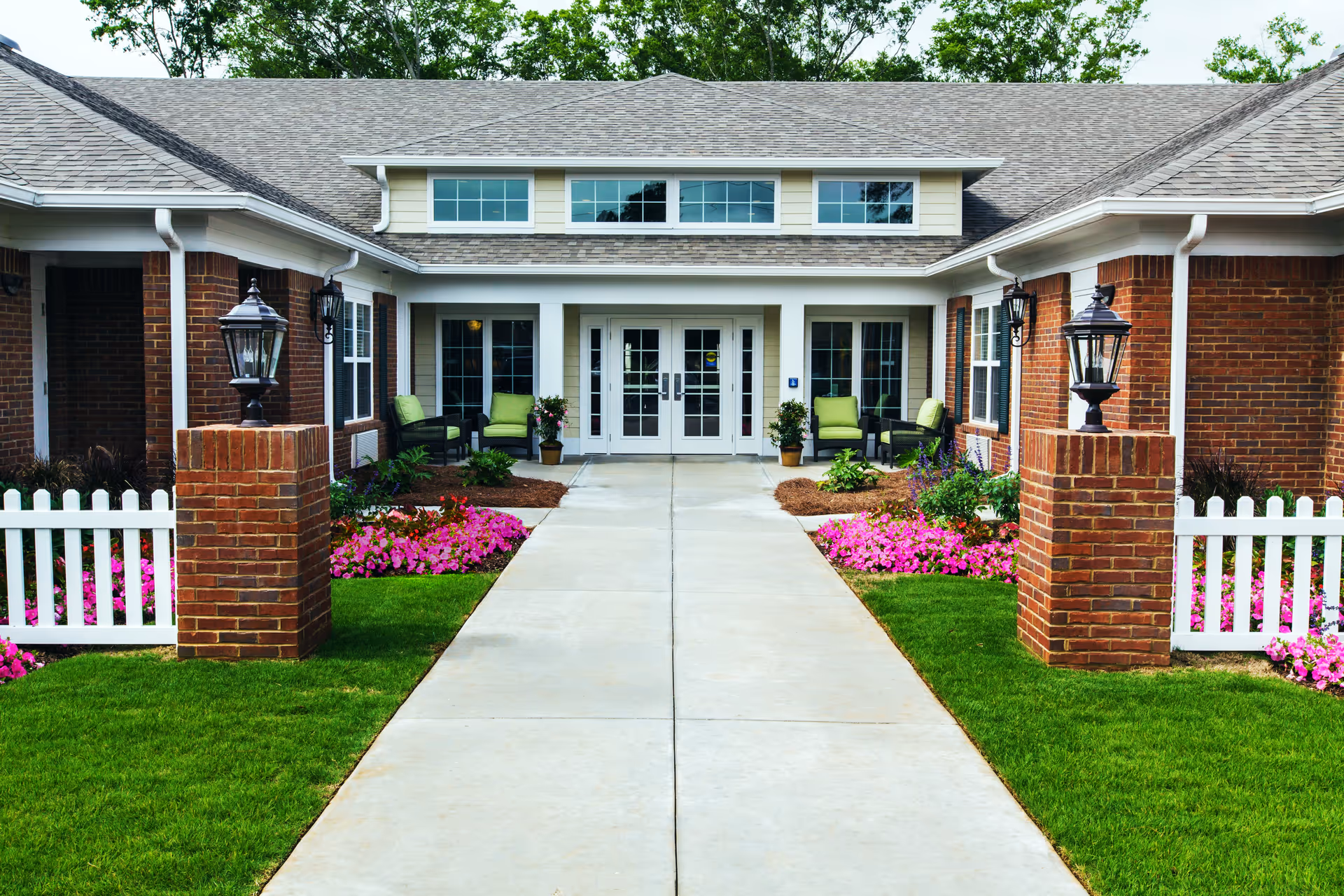 Front entrance of a senior living facility with a concrete walkway leading to double glass doors. The building has a brick exterior with white trim and a gray shingled roof. There are green cushioned chairs on either side of the entrance, surrounded by flower beds with pink and purple flowers and green shrubs. Two brick pillars with lantern-style lights flank the walkway, and white picket fences border the flower beds.
