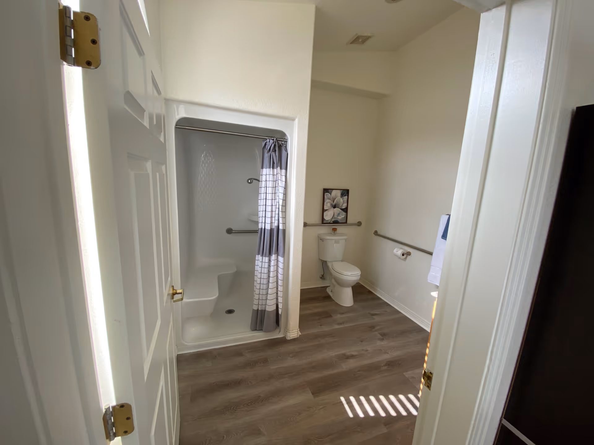 View into a bathroom with a white door partially open. Inside, there is a white shower with a built-in seat and a gray and white striped shower curtain. Next to the shower is a white toilet with a floral picture hanging above it. The bathroom has light-colored walls and wood-look flooring. There are metal grab bars on the walls near the toilet and shower for support.