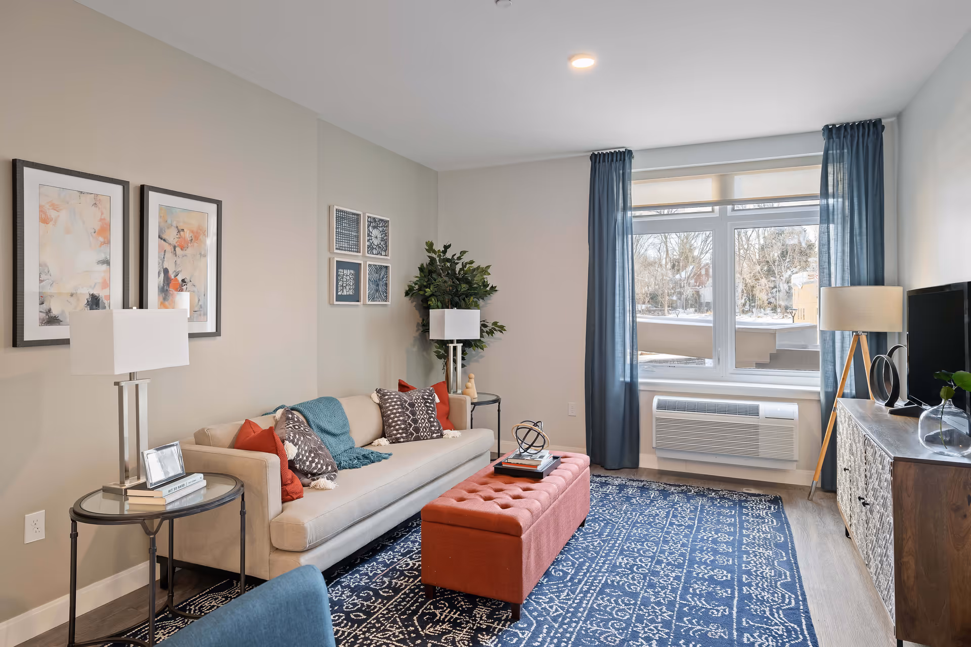 A cozy living room with a beige sofa adorned with patterned and solid red pillows, a blue throw blanket, and a red tufted ottoman in front. The room features a blue patterned rug, two framed abstract paintings on the wall, a side table with a lamp and books, a potted plant, a window with blue curtains, a floor lamp, and a TV on a wooden cabinet.