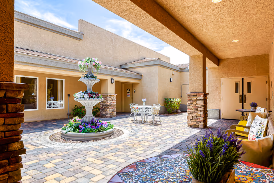 Outdoor courtyard area with a multi-tiered flower fountain in the center, surrounded by a paved stone floor. There are white metal chairs and tables, potted plants, and cushioned seating with decorative pillows under a covered patio. The building walls are beige with stone accents on the columns.