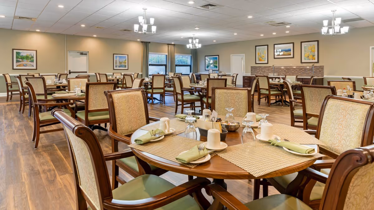 A spacious dining room with multiple wooden tables and chairs arranged neatly. Each table is set with placemats, napkins, cups, and glasses. The room has wooden flooring, soft lighting from ceiling chandeliers, and framed artwork on the walls.