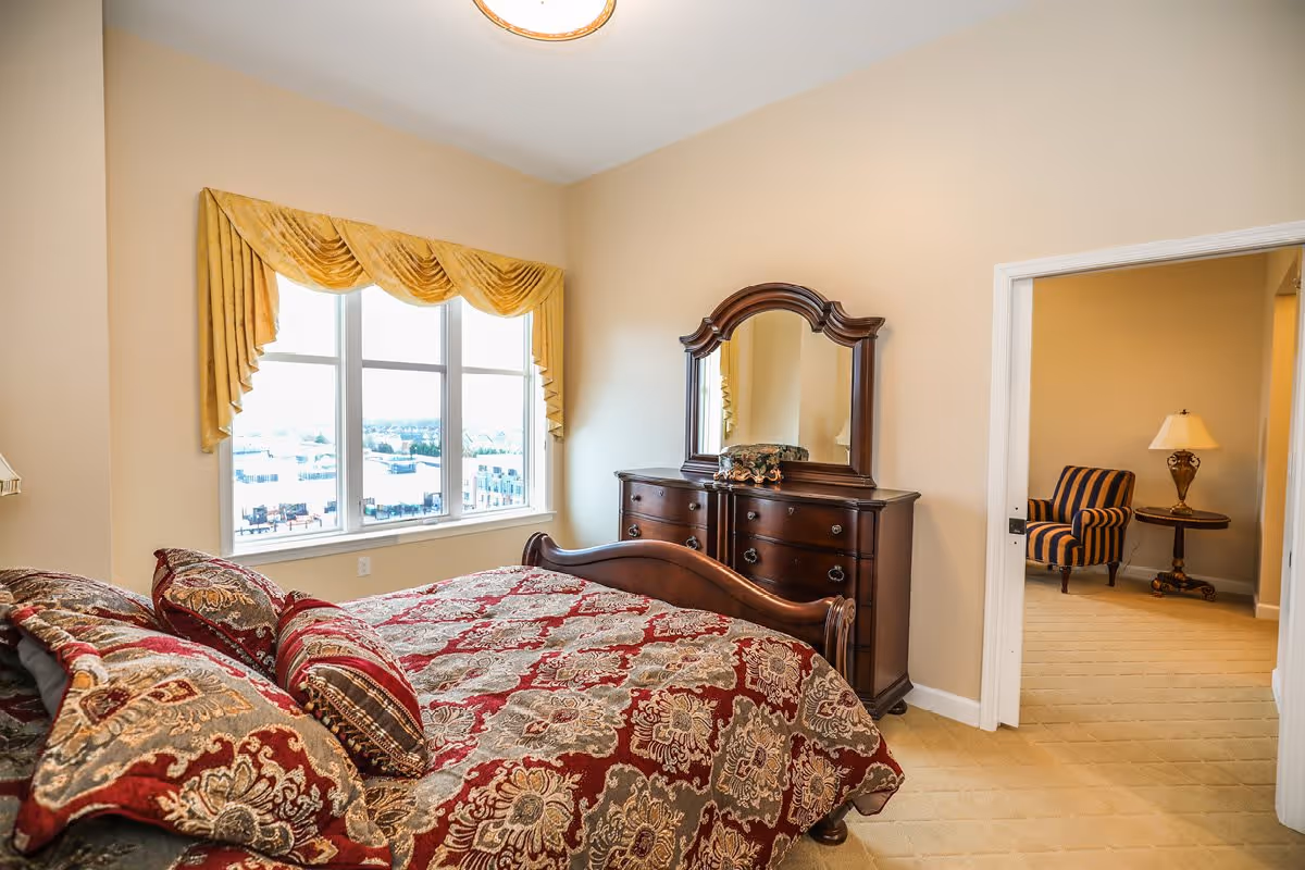 A bedroom with a large window covered by gold drapes, a wooden bed with a red and gold patterned bedspread and pillows, and a matching wooden dresser with a mirror. Through an open doorway, a room with a striped armchair, a side table, and a lamp is visible.