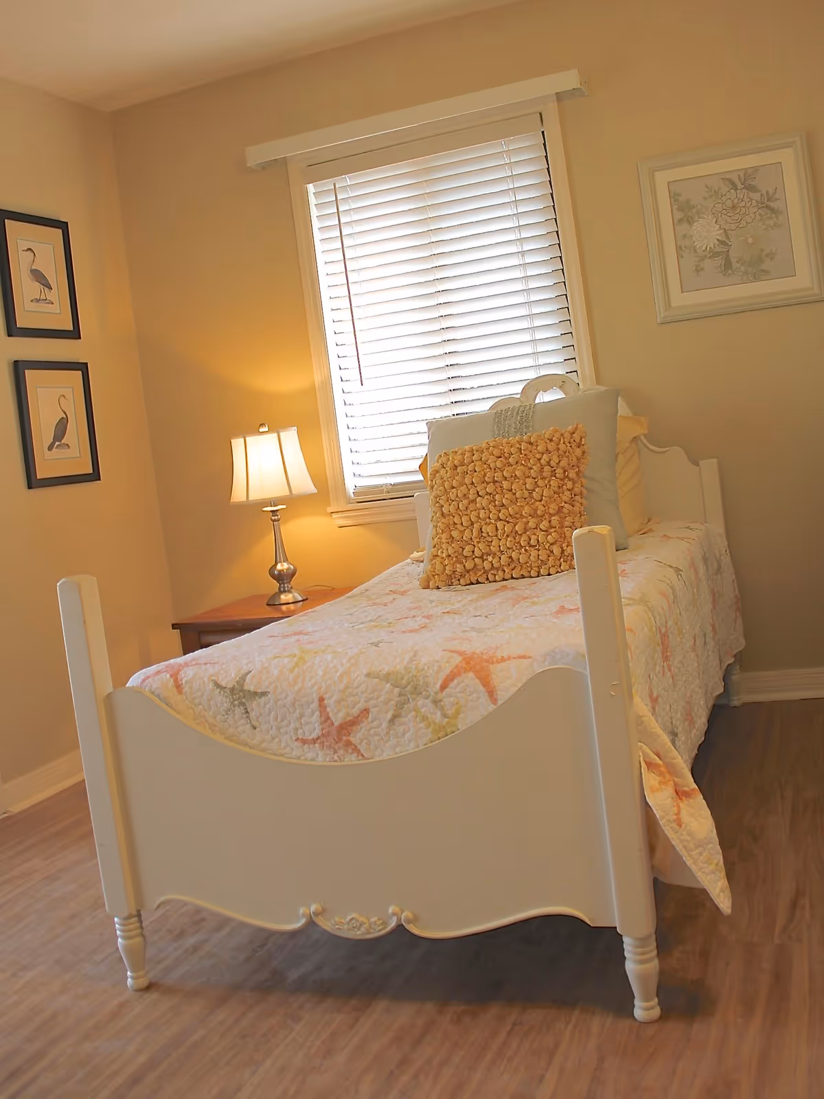 Warm, tidy single bedroom with a white wooden bed, patterned quilt and decorative pillows, a nightstand lamp, and framed artwork.