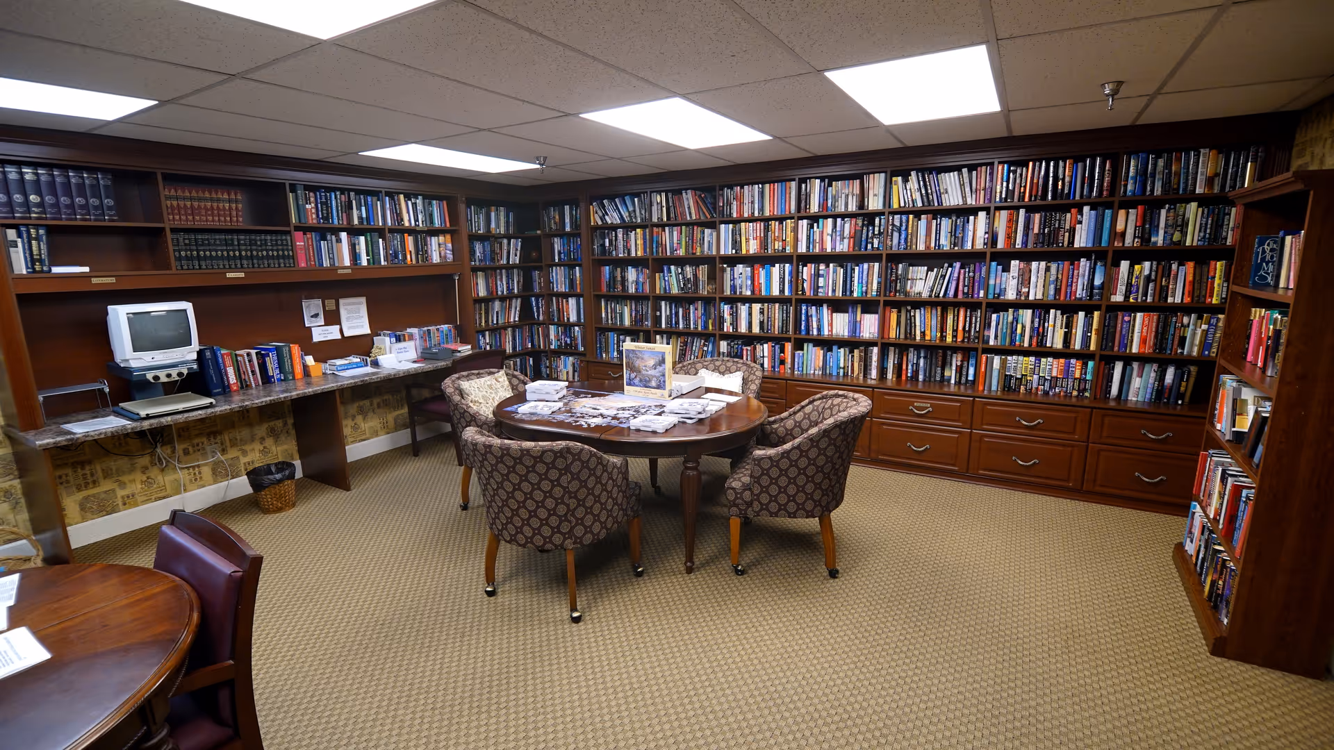 A cozy library room with walls lined with wooden bookshelves filled with books. In the center, there is a round wooden table surrounded by four patterned upholstered chairs. On the left side, there is a long counter with more books and an old computer monitor. The room has a beige carpet and a drop ceiling with fluorescent lighting.