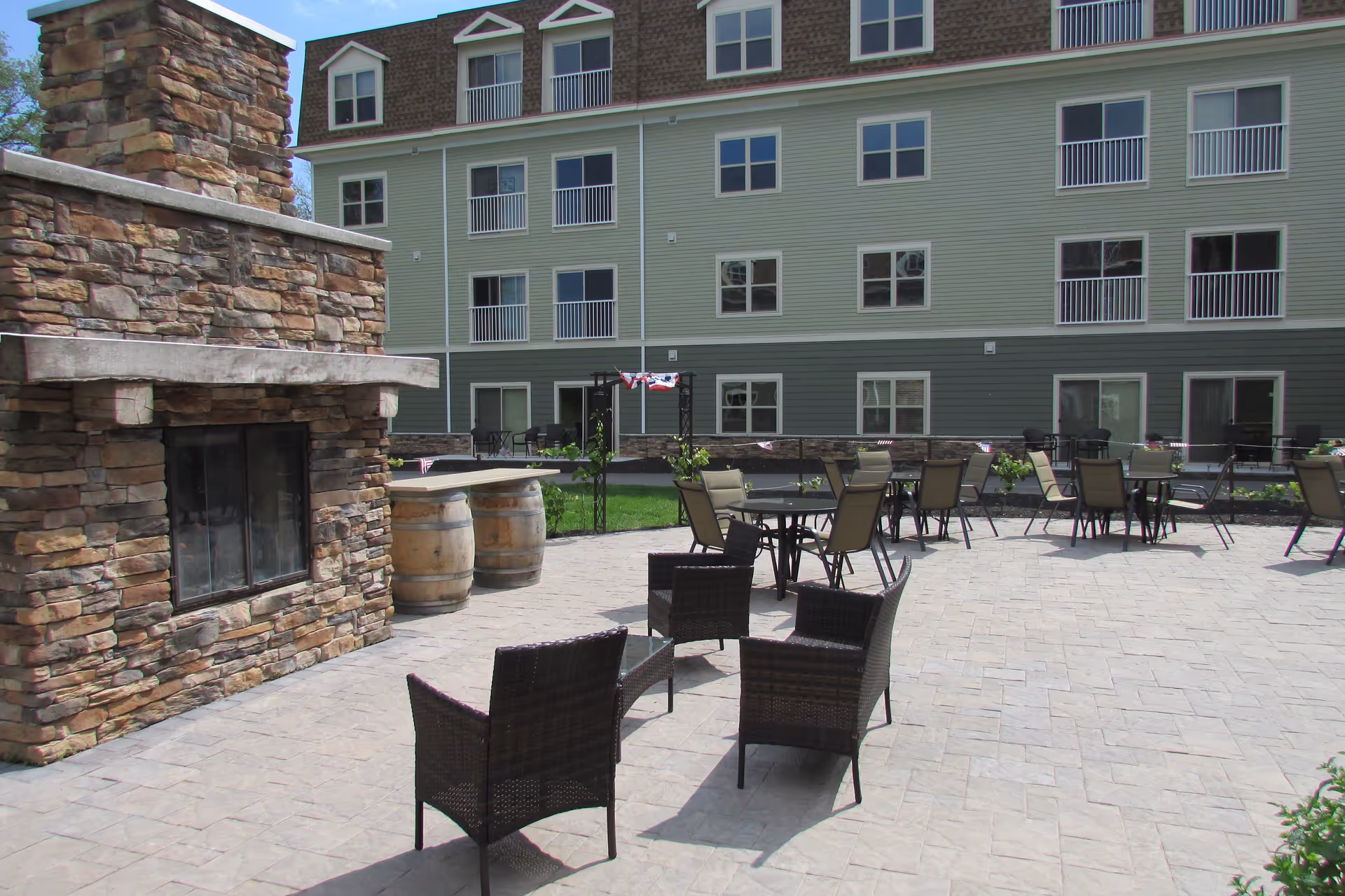 Outdoor patio area with stone fireplace, several tables and chairs, and a multi-story building in the background with multiple windows and balconies.