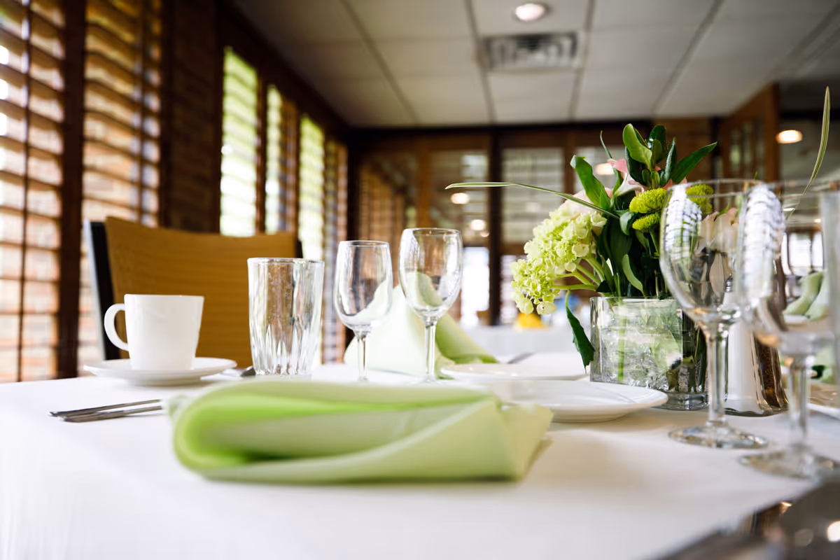 A dining table set with white tablecloth, green folded napkins, clear drinking glasses, wine glasses, white plates, silverware, and a vase with green and white flowers in a room with large windows and wooden blinds.