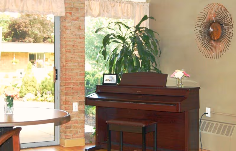 A cozy interior corner featuring a dark wooden piano with a matching bench, a large green plant behind it, and a small framed photo and vase with flowers on top. To the left, there is a round wooden table with a flower vase, and a sliding glass door with a view of greenery outside. A decorative sunburst mirror hangs on the beige wall to the right.