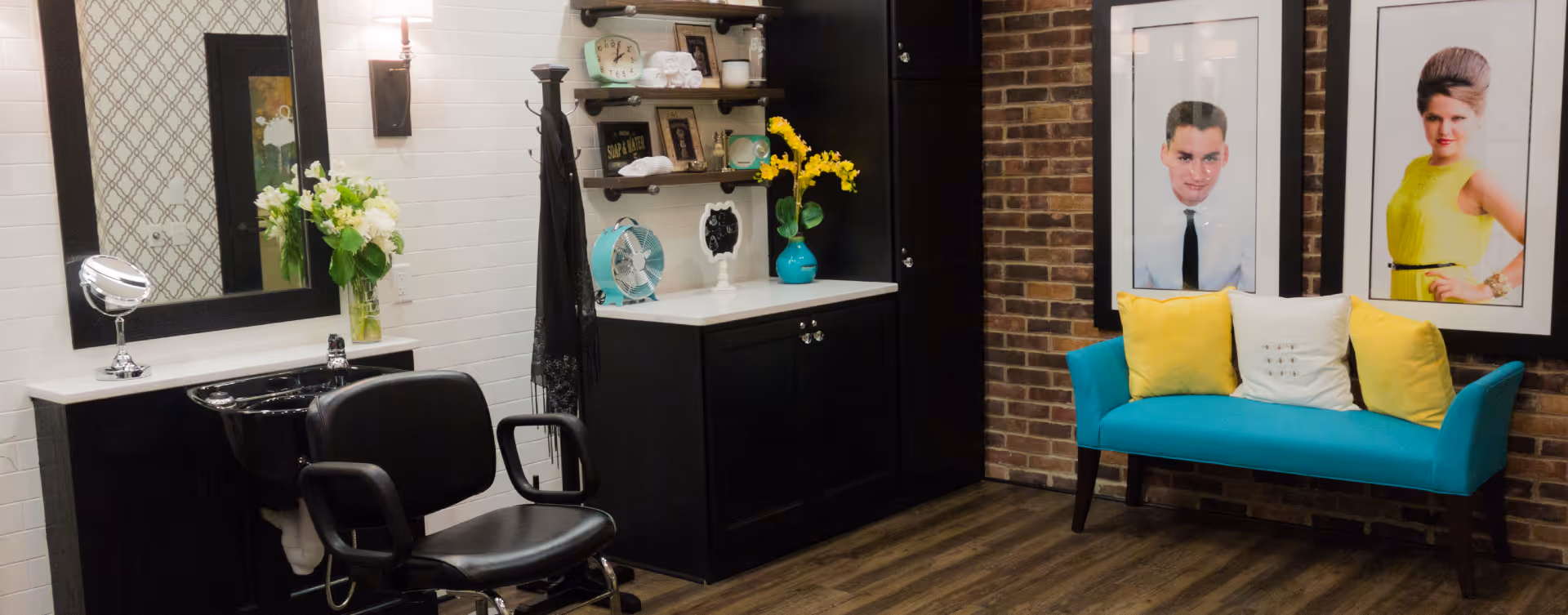 Interior of a salon area with a black salon chair and sink, a large mirror, a vase with flowers on the counter, shelves with towels and decor, a turquoise fan, and a turquoise bench with yellow and white pillows. Two large framed portraits hang on a brick wall behind the bench.