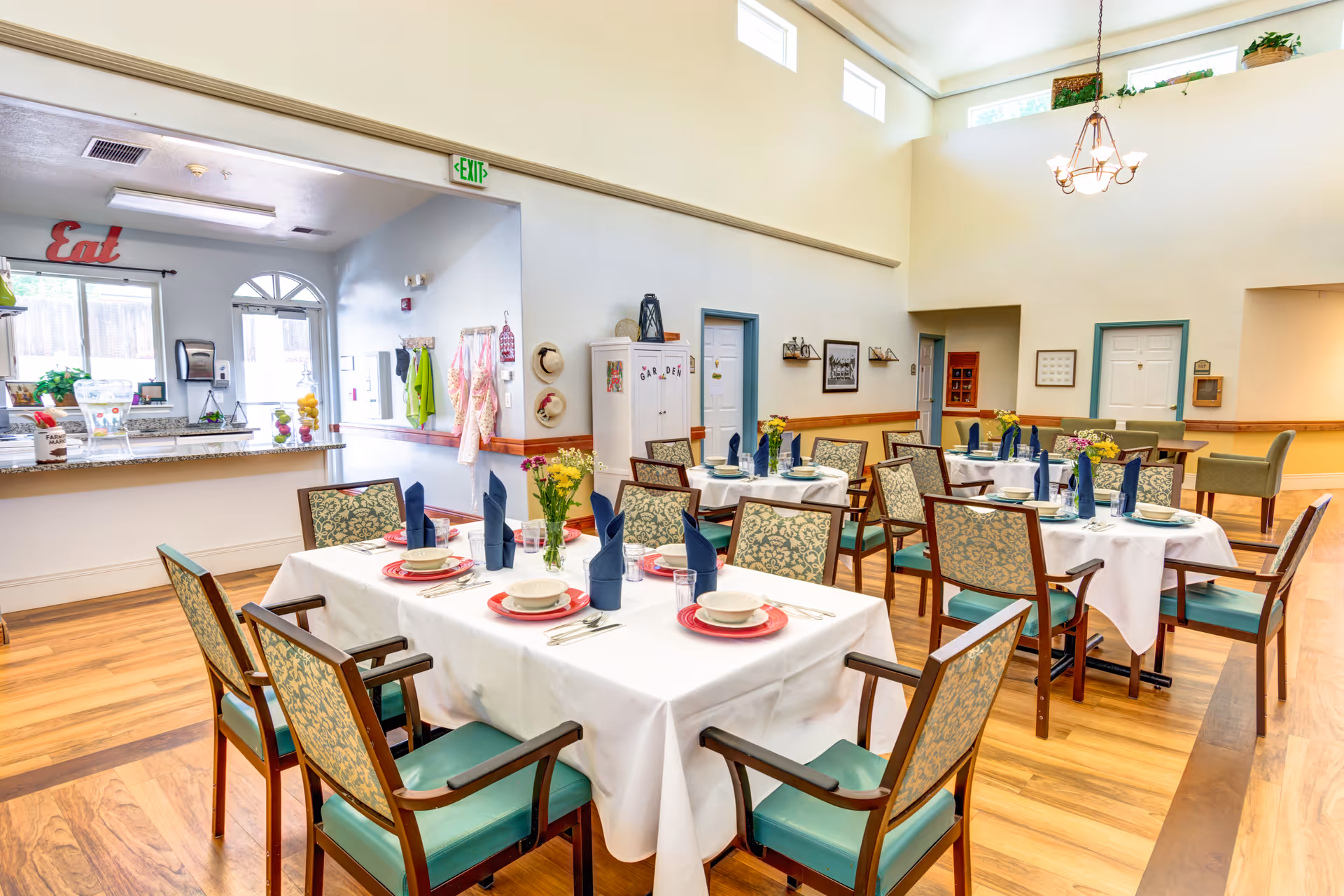 Bright dining room with several tables covered in white tablecloths, each set with plates, bowls, silverware, and blue folded napkins. The room has wooden floors, patterned chairs with green cushions, a chandelier, and high windows letting in natural light. A counter area with a sign that says 'Eat' is visible in the background.