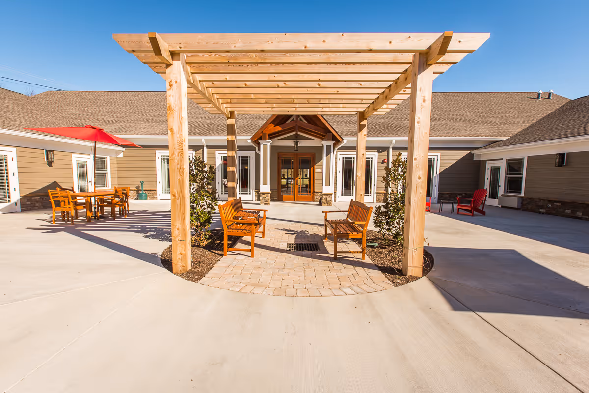 Outdoor courtyard area with a wooden pergola structure, two wooden benches underneath, and a paved walkway. Surrounding the pergola are small plants and a concrete patio with additional seating including a table with chairs and a red umbrella, as well as red Adirondack chairs near the building entrance. The building has beige siding, white trim, and a peaked roof with a covered entrance featuring double wooden doors.