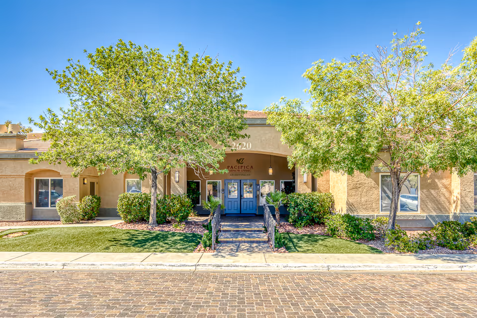 Front exterior view of a senior living facility named Pacifica Senior Living Green Valley, showing a beige building with blue double doors, surrounded by green trees and bushes under a clear blue sky.