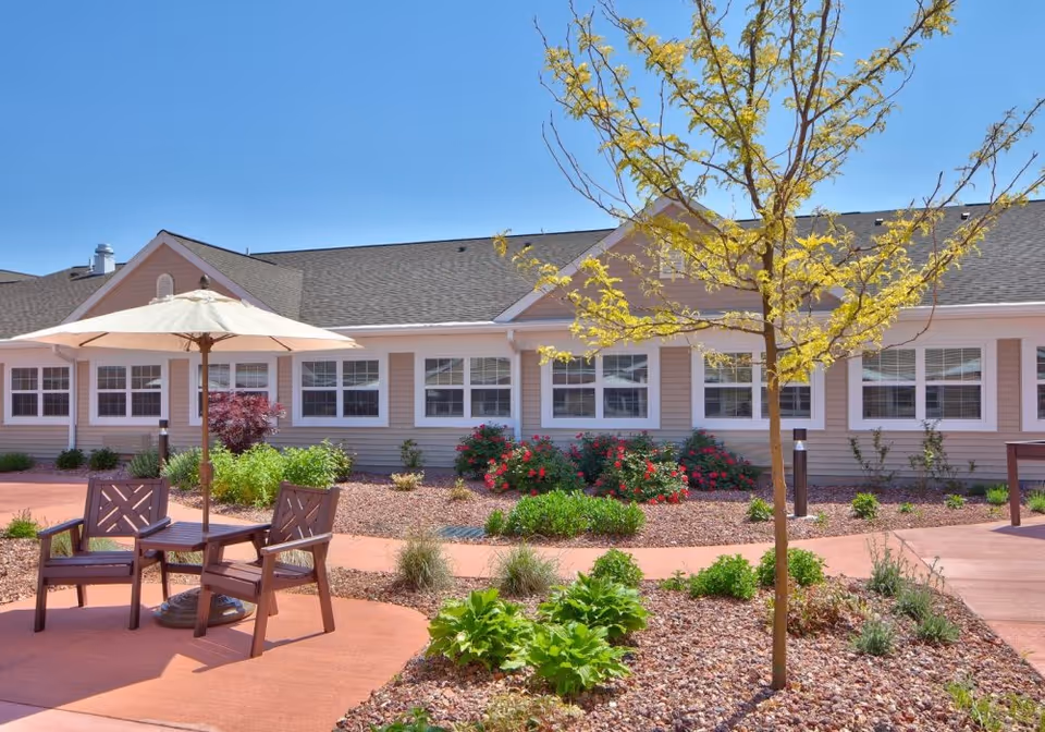 Outdoor patio area with a small table, two wooden chairs, and a large white umbrella. The patio is surrounded by landscaped garden beds with various plants and a young tree with yellow leaves. In the background, there is a single-story building with multiple windows and a gray roof under a clear blue sky.
