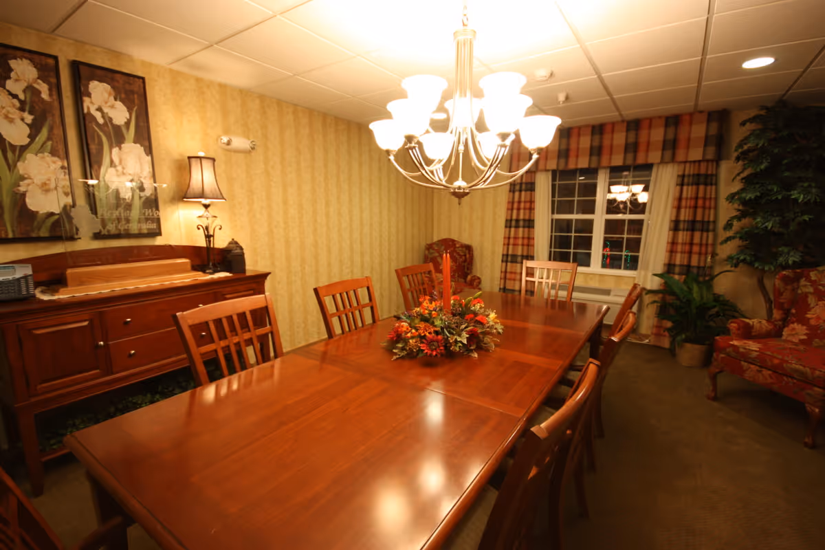 Well-lit dining room with a long wooden table and chairs, chandelier, sideboard, and a floral centerpiece.