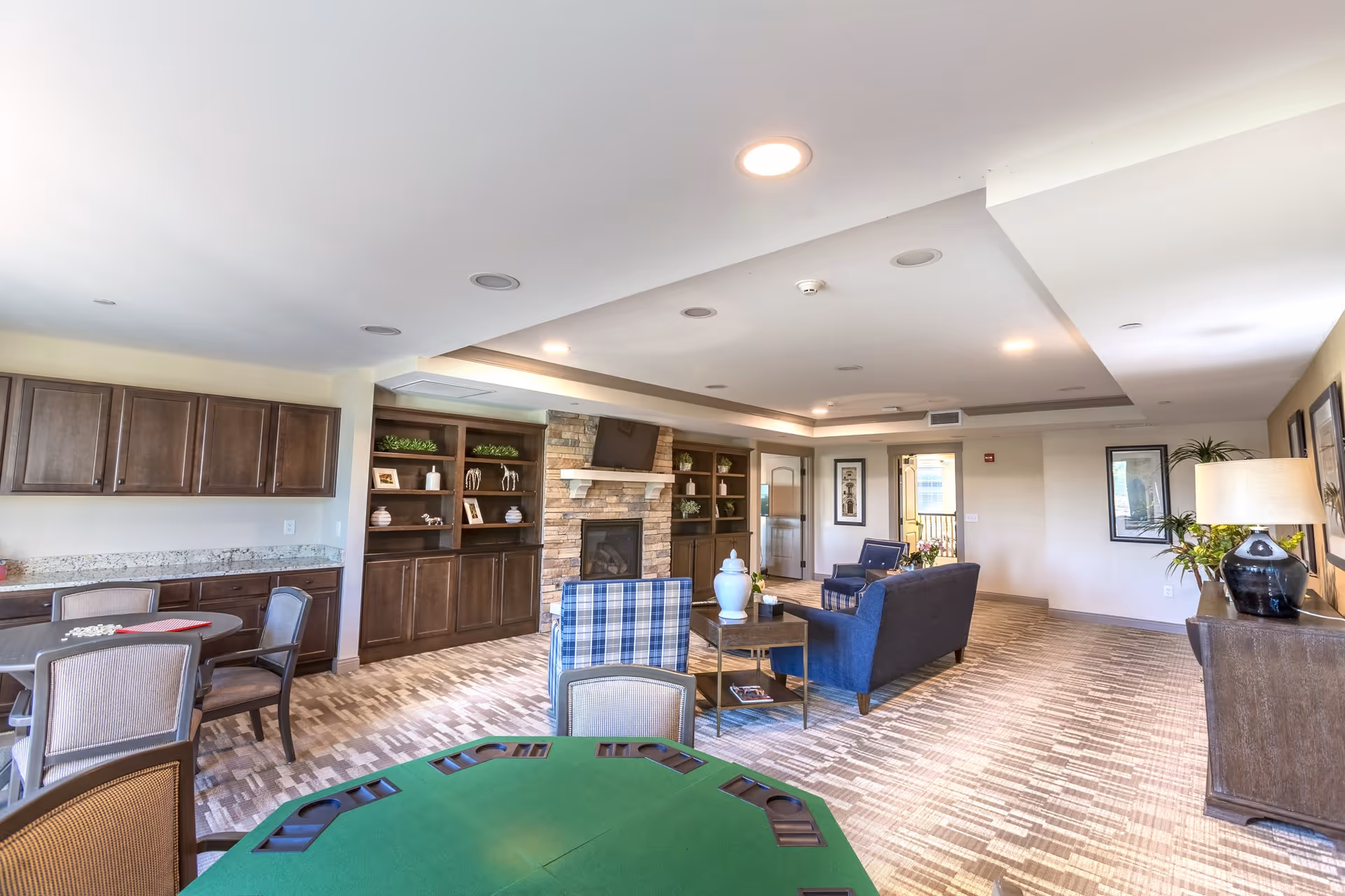 A spacious and well-lit common area in a senior living facility featuring a green card table in the foreground, several chairs, a seating area with blue armchairs and a sofa around a coffee table, built-in wooden shelves with decorative items, a stone fireplace with a mounted TV above it, and cabinets with a granite countertop along one wall. The room has carpeted flooring and recessed ceiling lights.