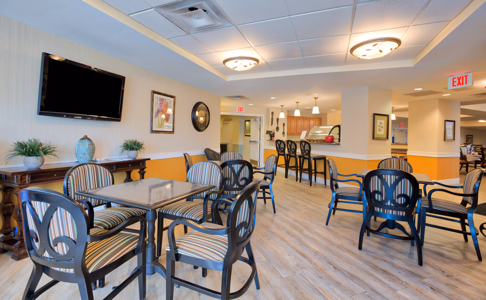 A bright and spacious common dining area in a senior living facility with multiple tables and striped cushioned chairs. The room features a wall-mounted flat-screen TV, decorative plants, framed artwork, and a clock on the wall. In the background, there is a counter with bar stools and pendant lighting, along with exit signs visible on the ceiling.