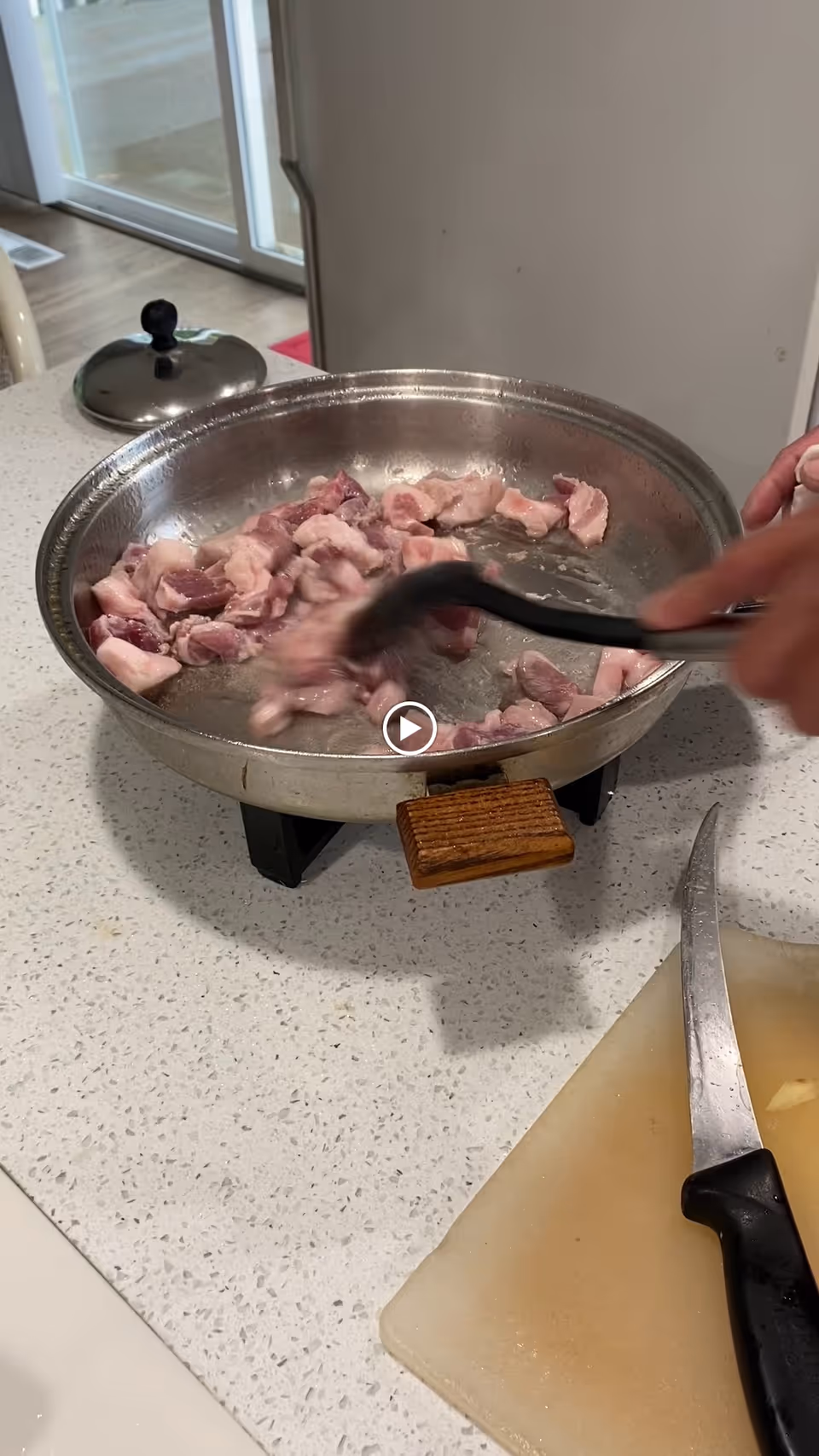A close-up view of raw meat pieces being stirred in a metal pan on a stovetop in a kitchen. A hand is holding a black spatula stirring the meat. A knife and a cutting board are visible on the countertop nearby.