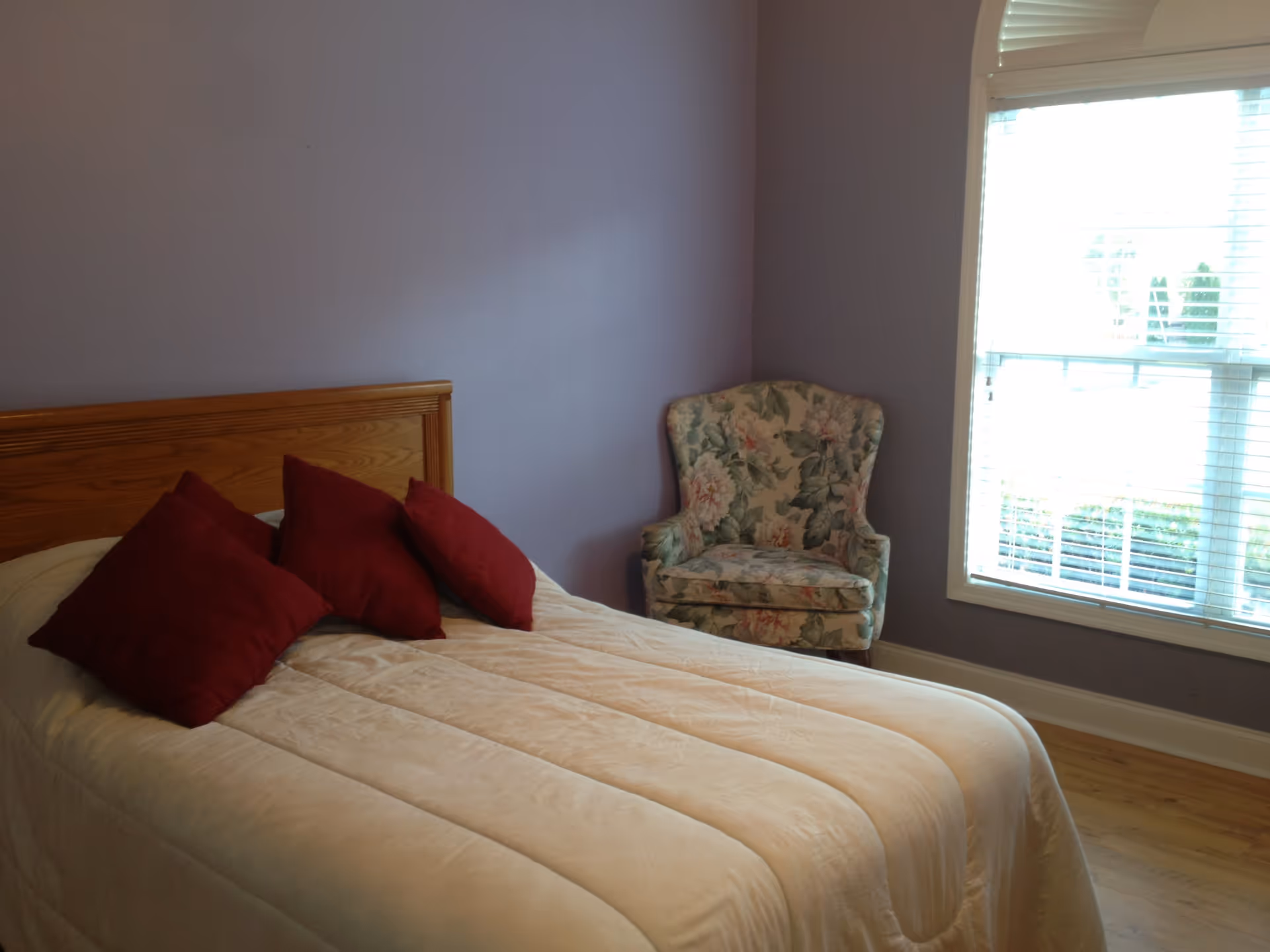 Small bedroom with a bed topped by three red pillows, a floral armchair, and a window with blinds against lavender walls.