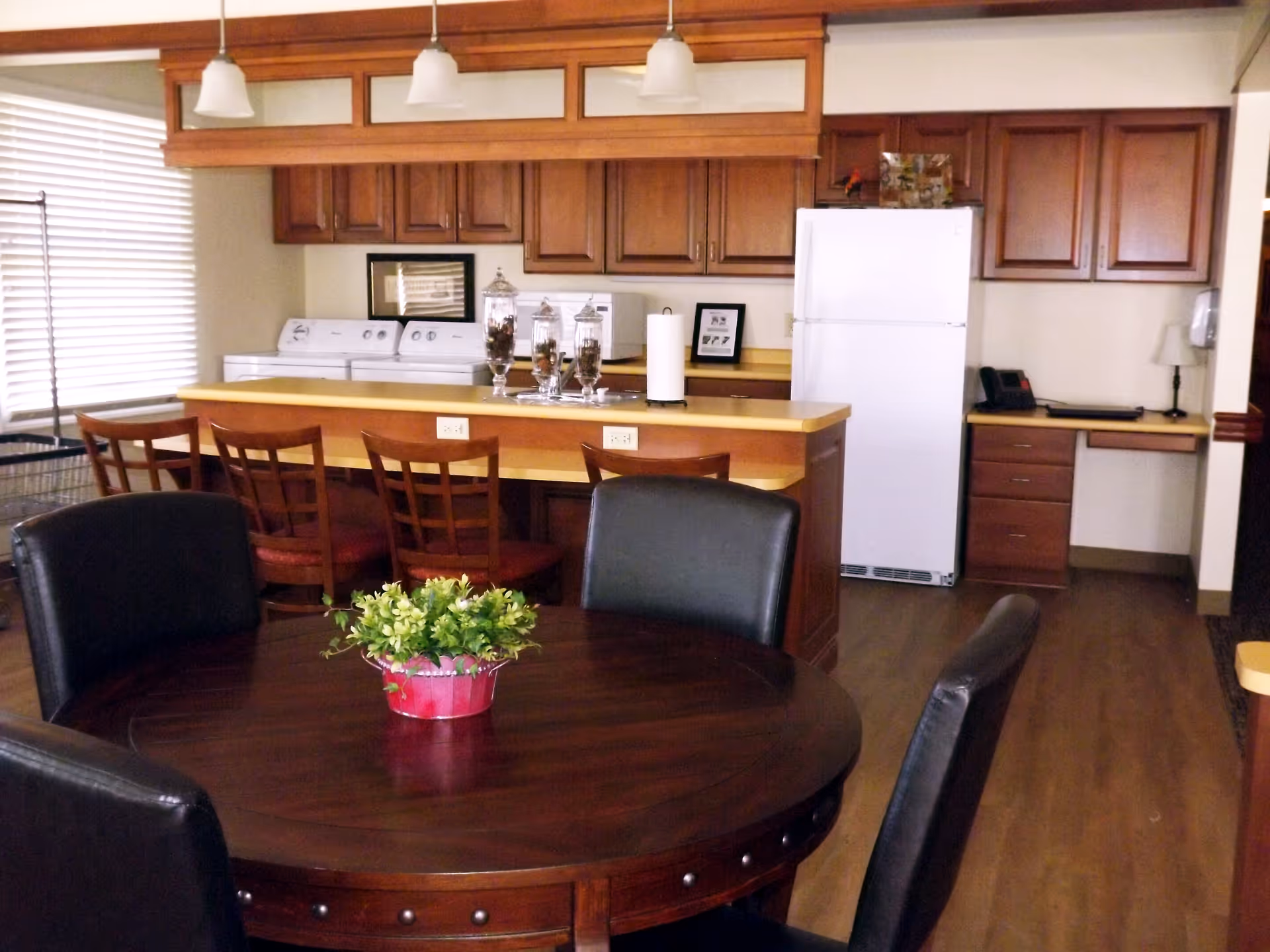 Interior view of a kitchen and dining area featuring a round wooden dining table with black chairs and a small plant centerpiece. The kitchen has wooden cabinets, a white refrigerator, a countertop with bar stools, and a washing machine and dryer near a window with blinds.