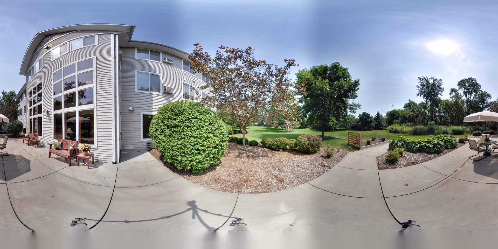 Outdoor patio and landscaped grounds beside a modern senior living building with large windows, benches, umbrellas, and walking paths.