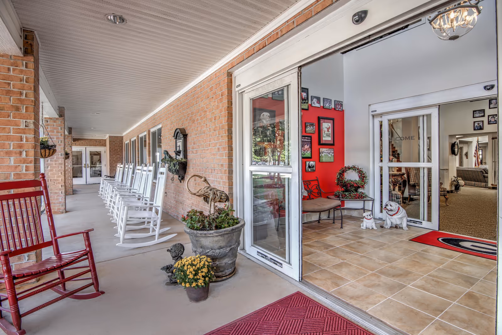 Covered outdoor porch area with a row of white rocking chairs along a brick wall and a single red rocking chair in the foreground. The porch leads to an entrance with glass doors opening into a tiled lobby area decorated with framed pictures on a red accent wall, a bench, and two dog statues. There are potted plants and a red welcome mat near the entrance.