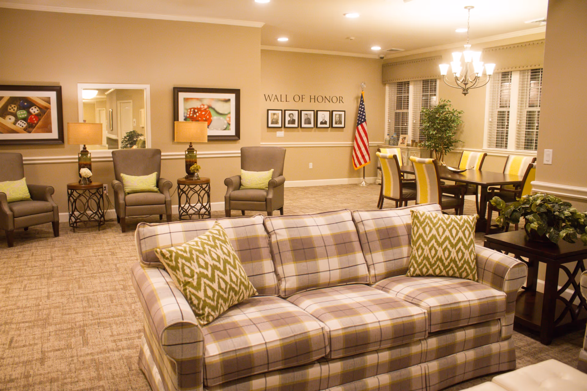 A cozy and well-lit common area in a senior living facility featuring a plaid sofa with green patterned pillows in the foreground, several armchairs with side tables and lamps along the back wall, a dining table with yellow and white chairs near windows, a 'Wall of Honor' with framed photos, and an American flag in the corner.