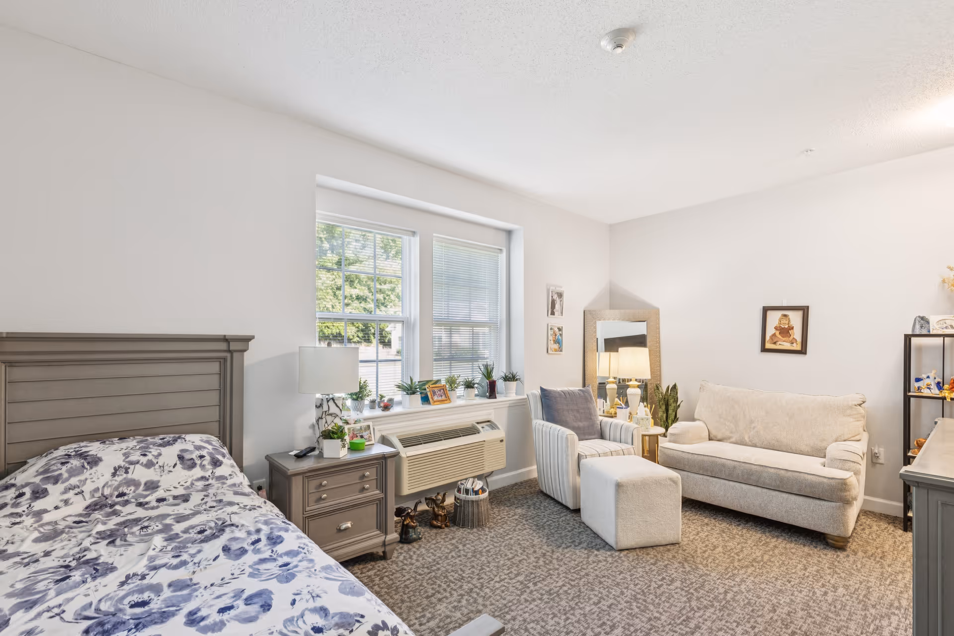 A cozy senior living room and bedroom area featuring a bed with floral bedding, a nightstand with a lamp and plants, a window with blinds, a striped armchair with a cushion, a small ottoman, a beige sofa, a tall mirror, and a shelving unit with decorative items. The room has light-colored walls and carpeted flooring.