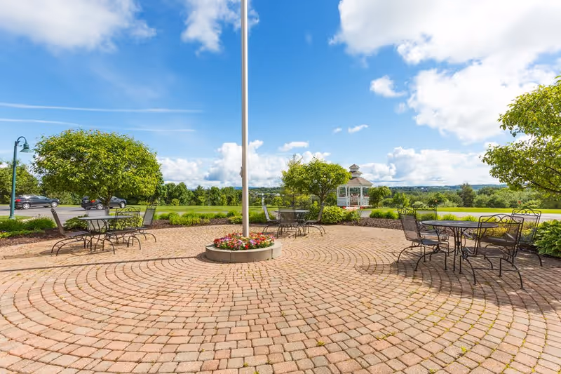 Outdoor patio area with circular brick paving, metal tables and chairs, small trees, and a white gazebo in the background under a partly cloudy blue sky.