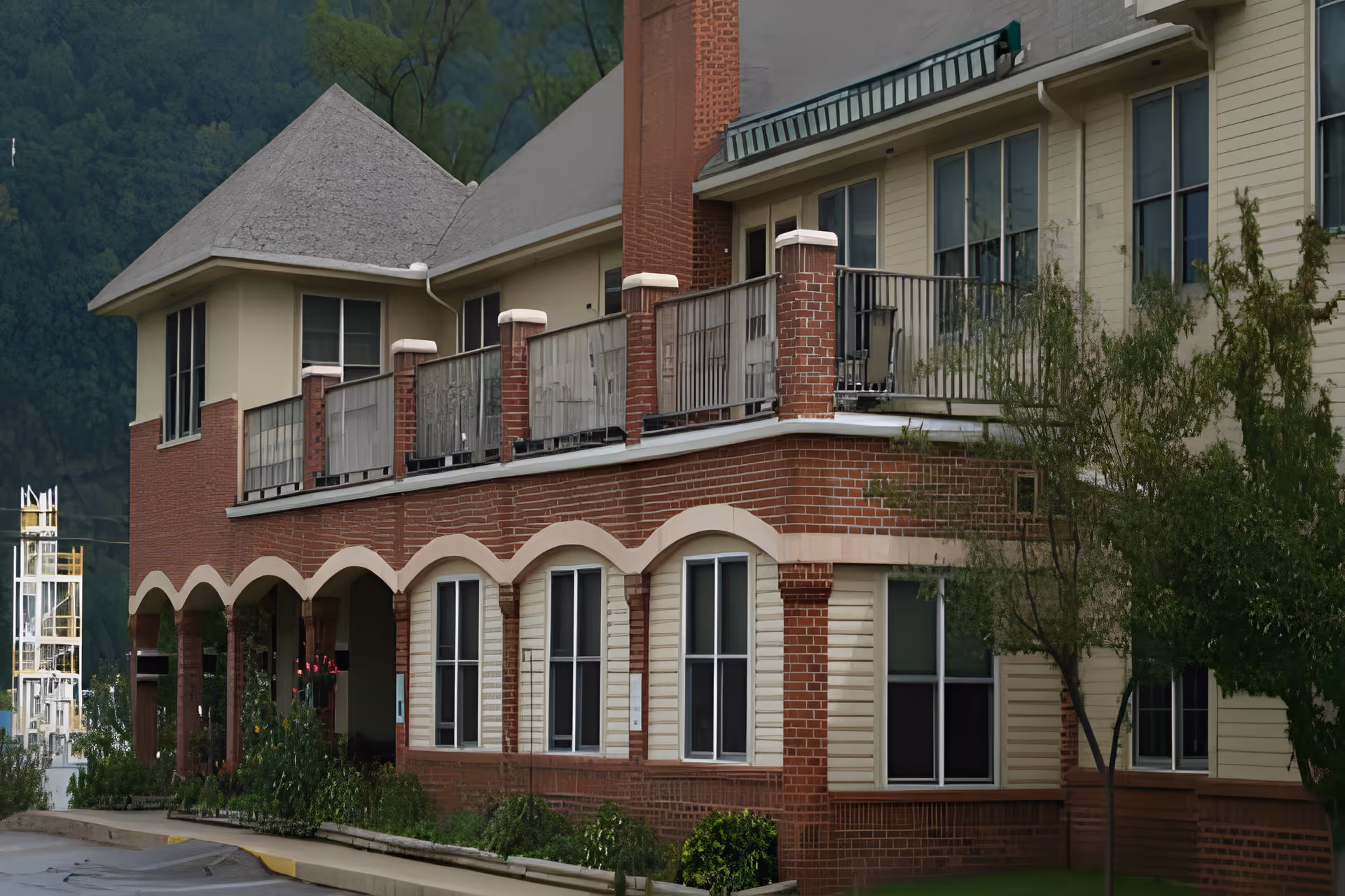 Exterior view of a two-story building with a brick and beige siding facade, featuring multiple windows, a balcony with railing on the second floor, and a covered walkway with arches on the ground floor. There are some trees and shrubs near the building and a mountain landscape in the background.