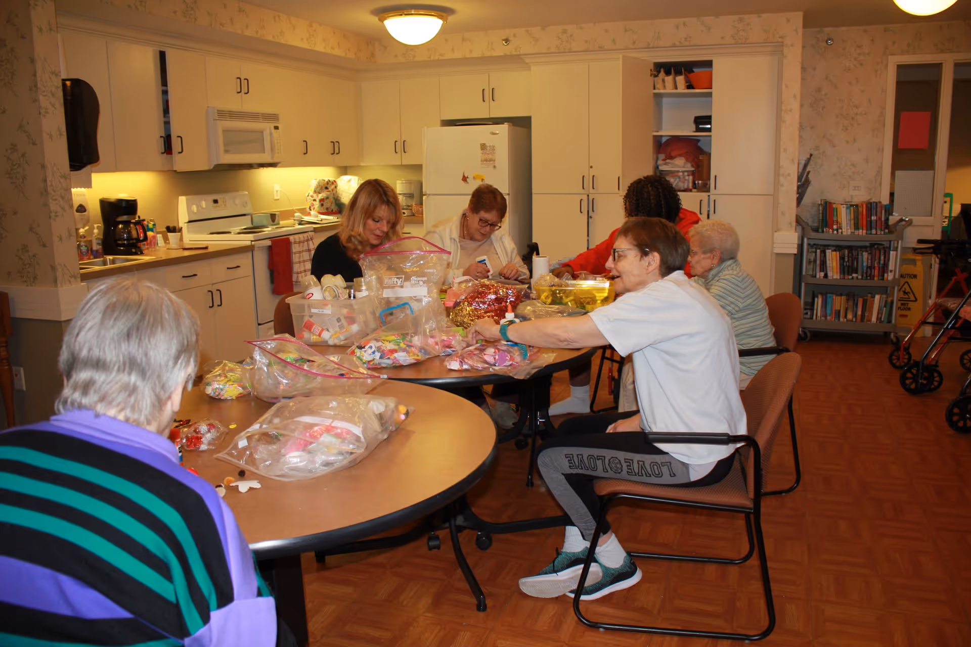 A group of elderly people and a caregiver sitting around two tables in a kitchen area, sorting and organizing items in plastic bags. The kitchen has white cabinets, a refrigerator, a microwave, and a coffee maker. There is a bookshelf and a walker visible in the background.