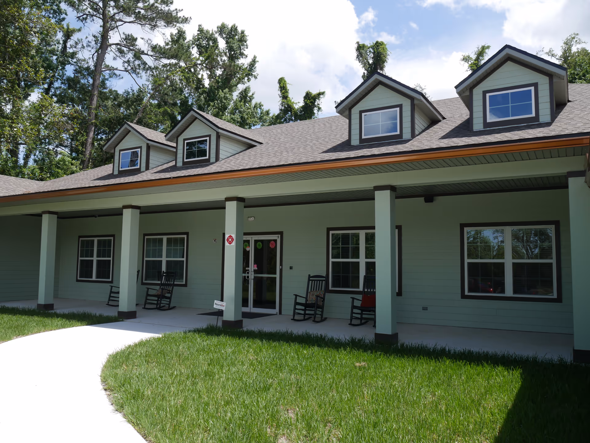 Exterior view of a single-story building with a covered porch featuring rocking chairs. The building has a gray roof with dormer windows and light green siding. There is a concrete walkway leading up to the porch, and green grass in the foreground with trees in the background under a partly cloudy sky.