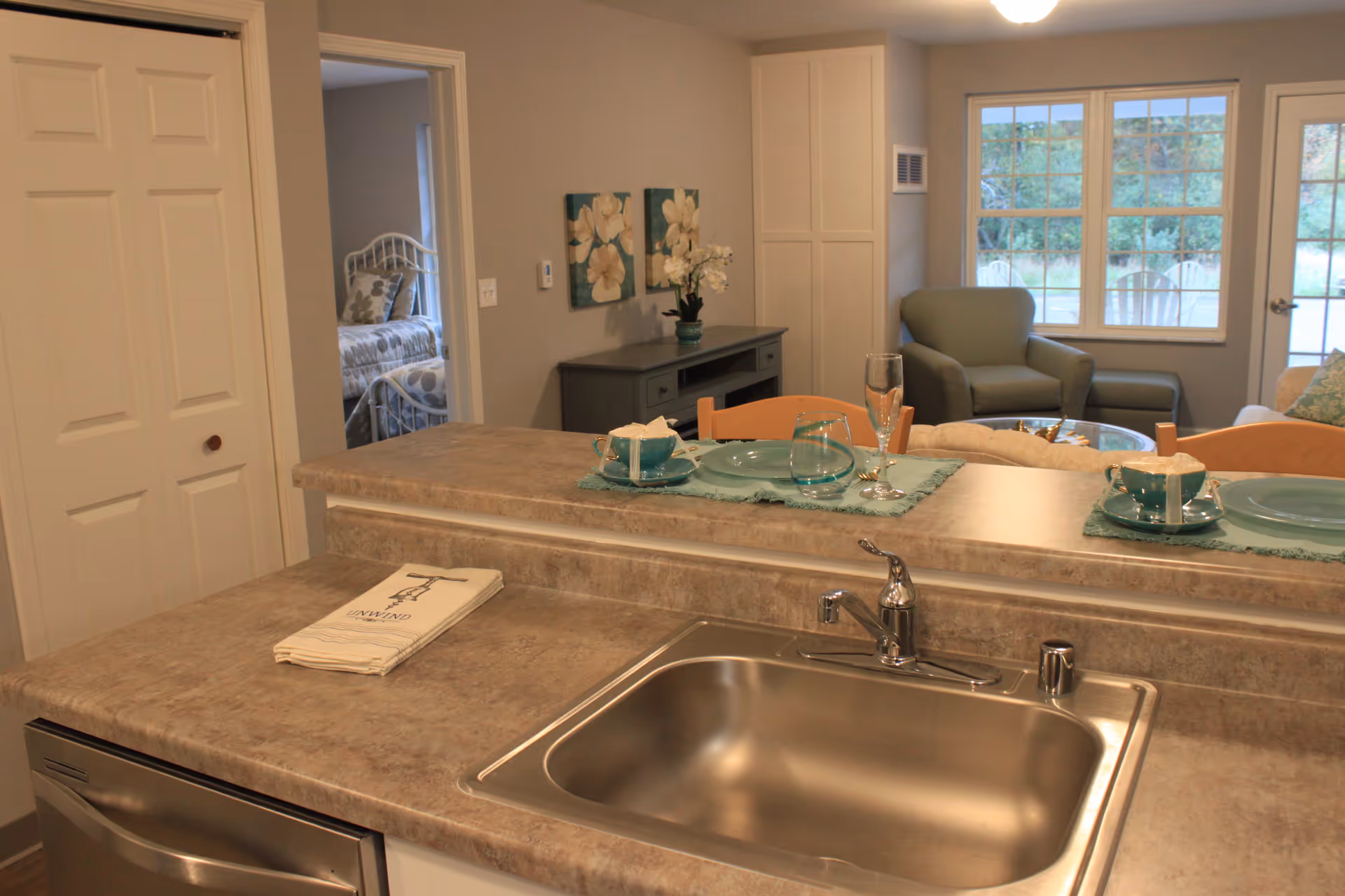 Kitchen countertop with a stainless steel sink and place settings overlooking a cozy living area and a bedroom doorway.