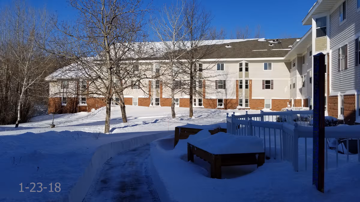Snow-covered outdoor area of a senior living facility with a curved walkway, leafless trees, and a multi-story building with white siding and brick accents under a clear blue sky.