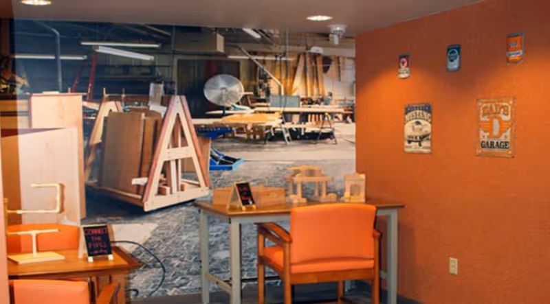 A small room with orange walls and orange chairs around wooden tables. On the tables are small wooden craft projects. The back wall features a large mural of a workshop with woodworking tools and equipment. Several vintage-style metal signs are mounted on the orange wall.